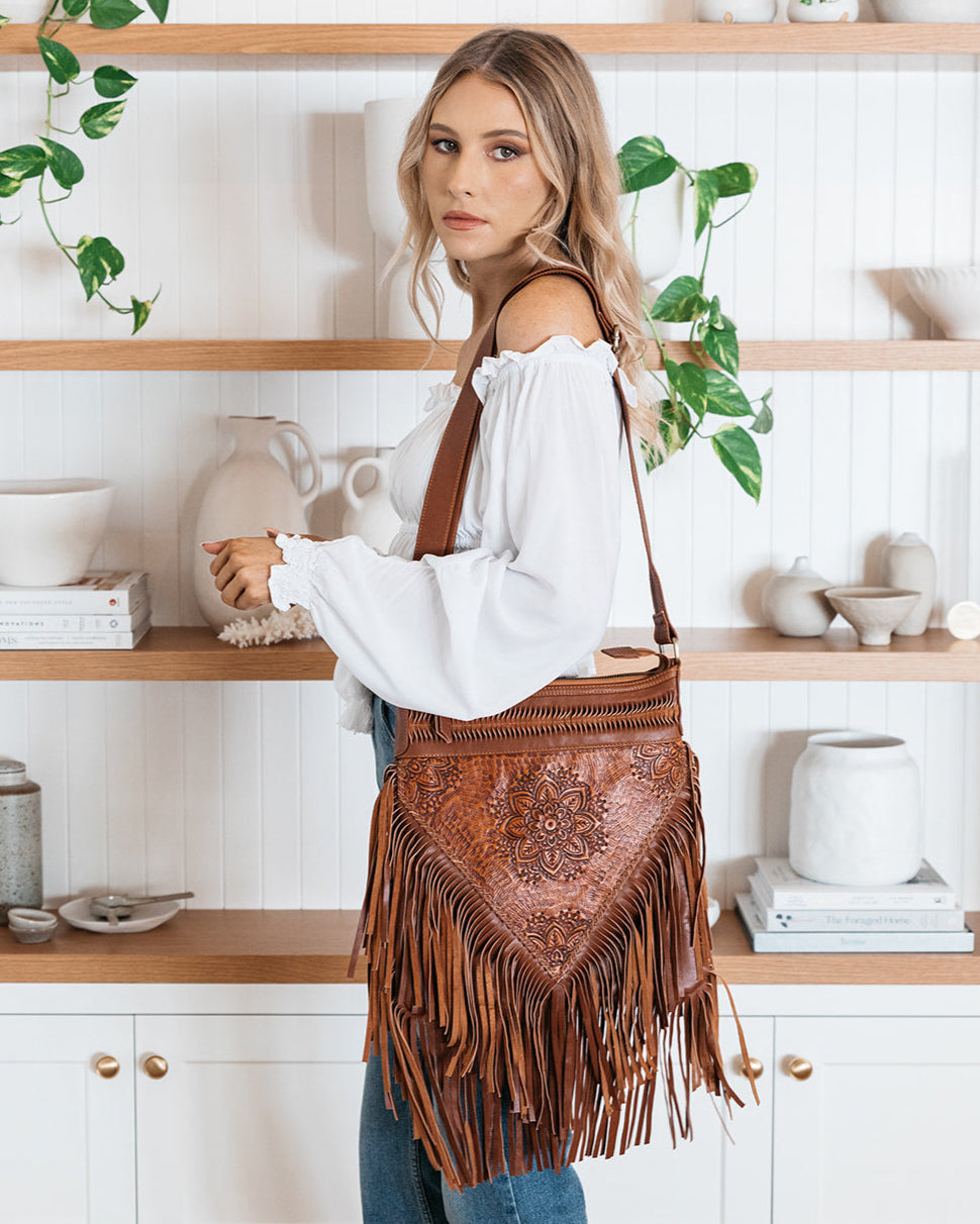Woman holding a brown leather bag with fringe in a home setting.