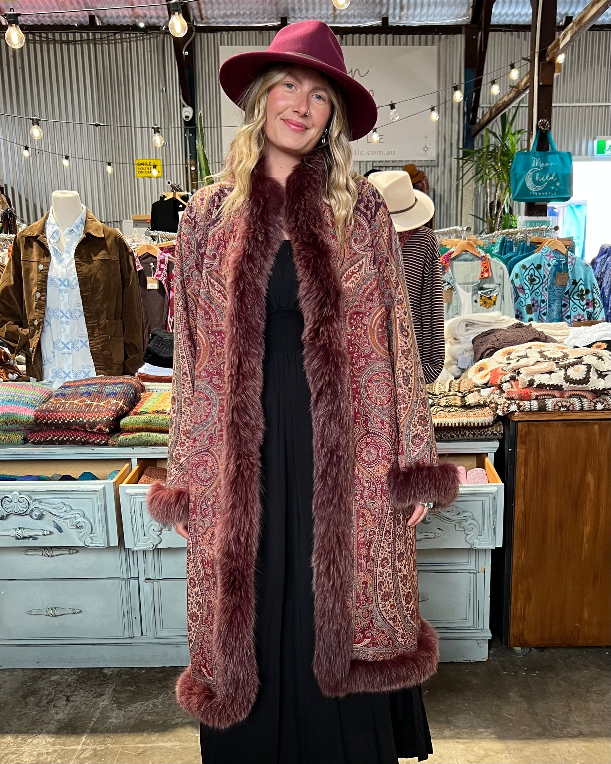A woman wearing a long burgundy faux fur coat with a paisley pattern, a matching wide-brim hat, and a black dress, standing in a vintage clothing store.