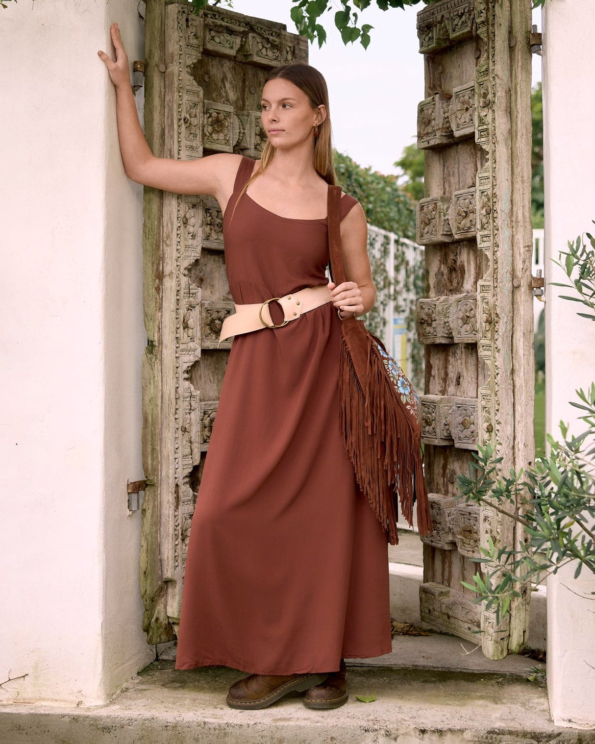 A woman stands in an open stone doorway framed by white walls and lush green foliage, wearing a rust-brown sleeveless maxi dress with a wide beige belt and carrying a matching fringed shoulder bag.