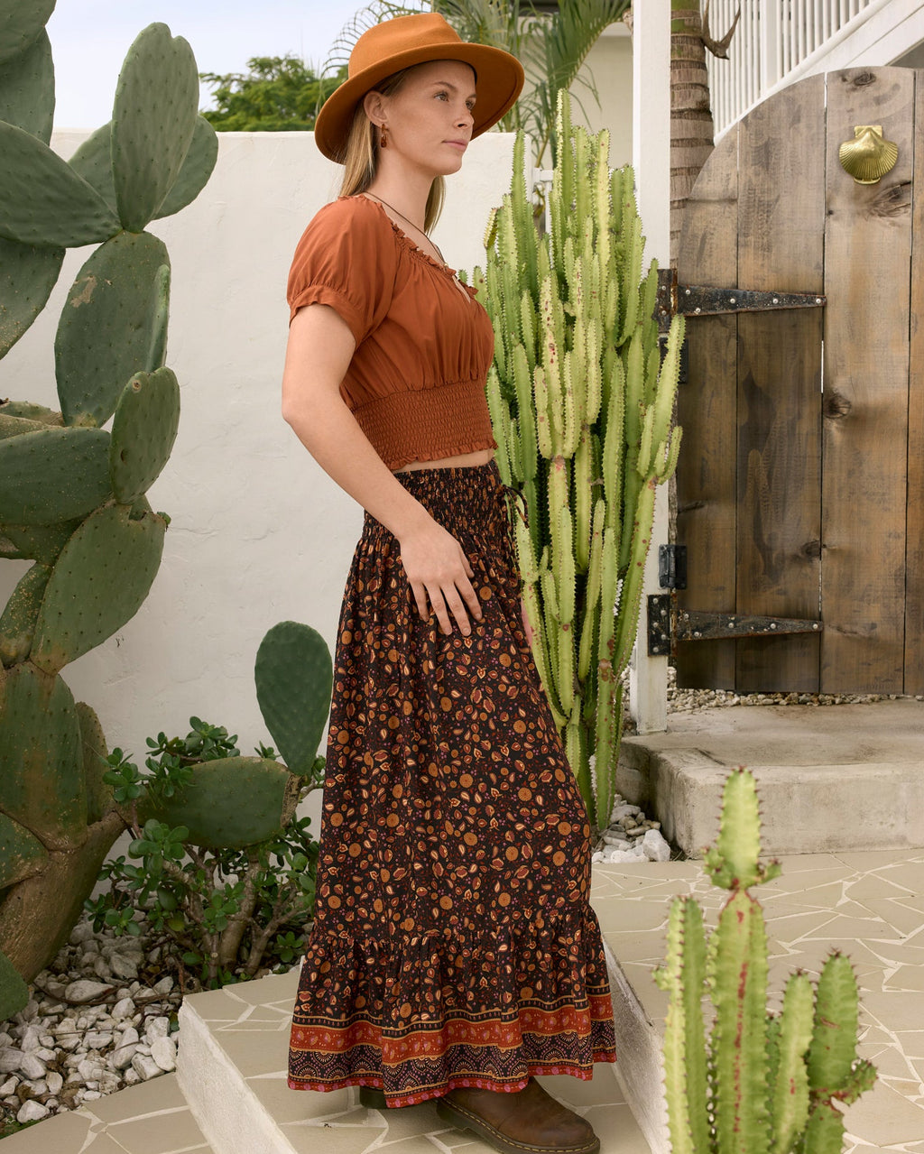 A woman stands outdoors in a desert garden setting wearing ava top gingerbread sleeve crop top, a dark brown bohemian floral maxi skirt, la luna and a wide-brimmed tan hat side veiw