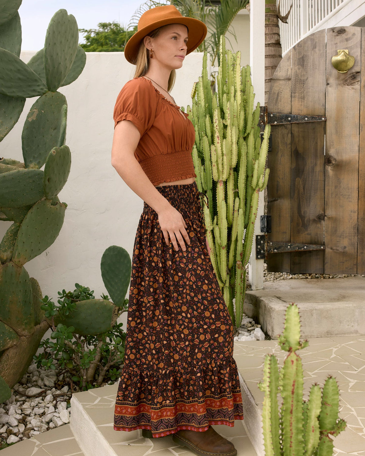 A woman stands outdoors in a desert garden setting wearing ava top gingerbread sleeve crop top, a dark brown bohemian floral maxi skirt, la luna and a wide-brimmed tan hat side veiw