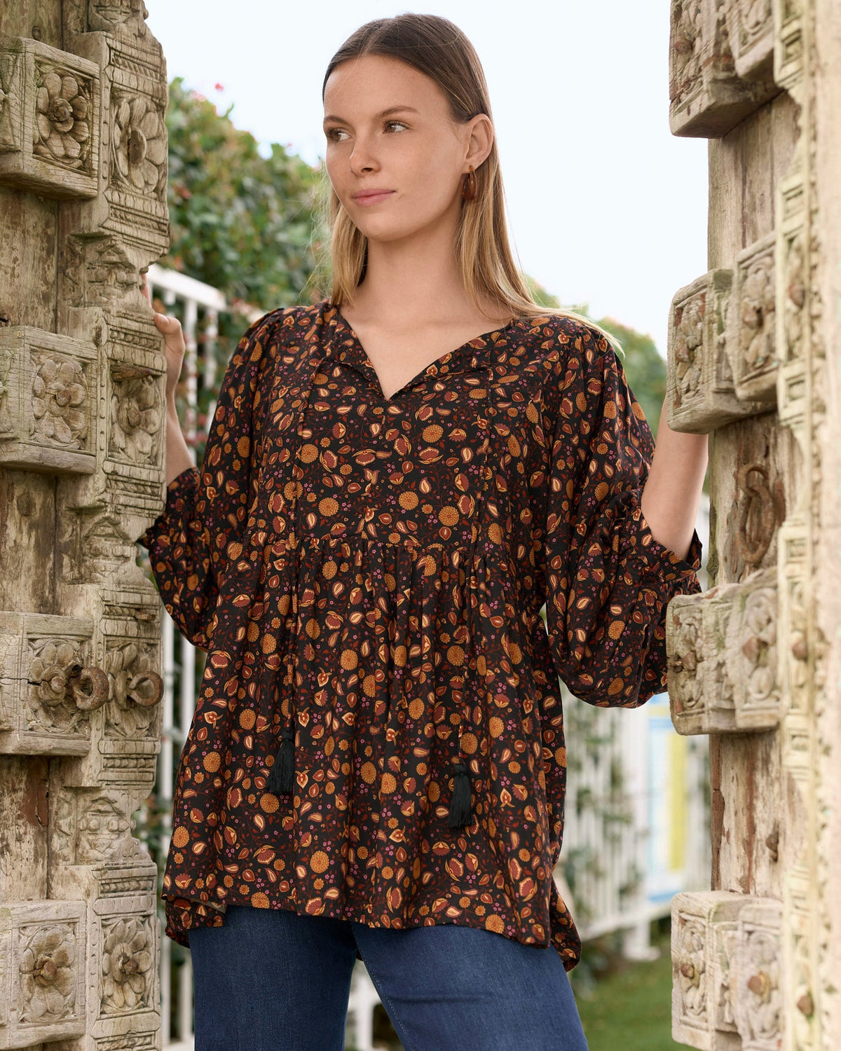 A woman wearing la luna print brown and orange boho patterned bambie v neckline top with tassels with blue jeans