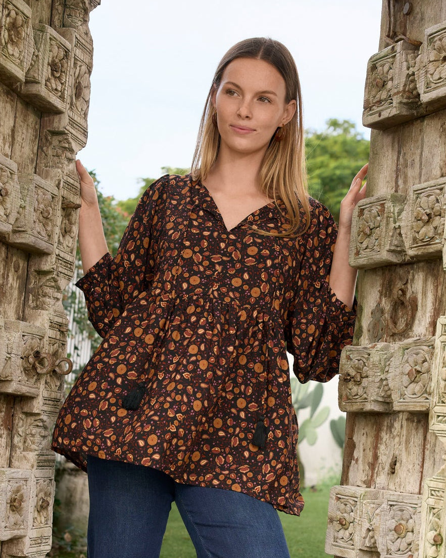 A woman modeling a brown short-sleeve button-up blouse with a small orange and yellow floral pattern, paired with blue jeans, standing in an open stone doorway.