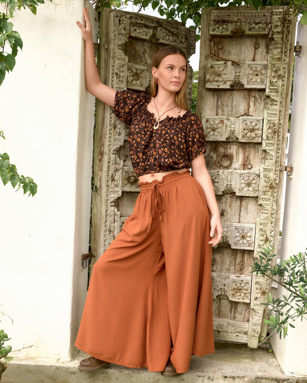 A woman stands in front of an ornate weathered wooden door framed by green foliage, wearing kia top la luna black base earthy floral short-sleeve crop top and high-waisted gingerbread billy pant wide-leg pants.