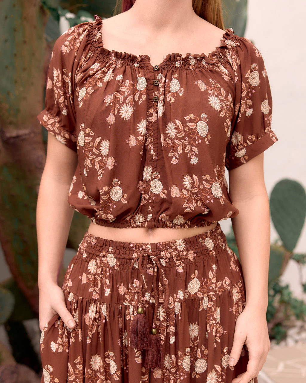 woman wearing ruched elastic neckline, soft elastic at hemline kai crop top with cuffed sleeve, brass buttons at neckline in bohemian brown floral dahlila print close up front view