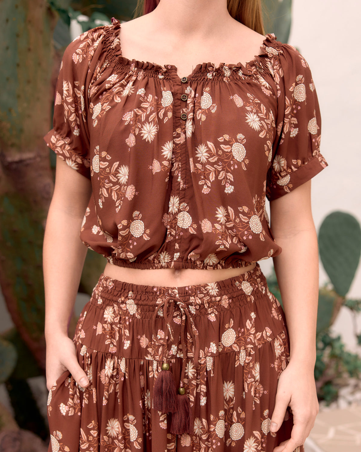 woman wearing ruched elastic neckline, soft elastic at hemline kai crop top with cuffed sleeve, brass buttons at neckline in bohemian brown floral dahlila print close up front view