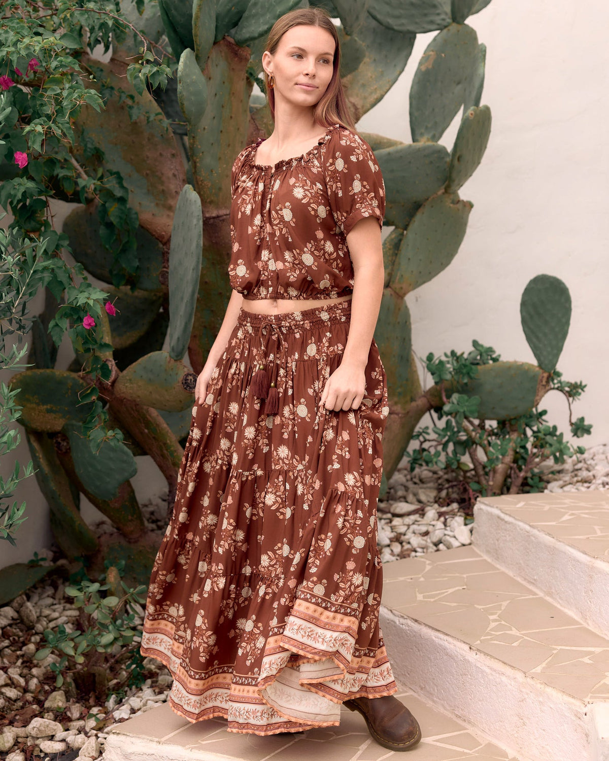 A woman modeling a brown bohemian-style two-piece set featuring a short-sleeve top and a matching maxi skirt with a cream floral print and lace hem, set against a background of cacti and outdoor stone steps.