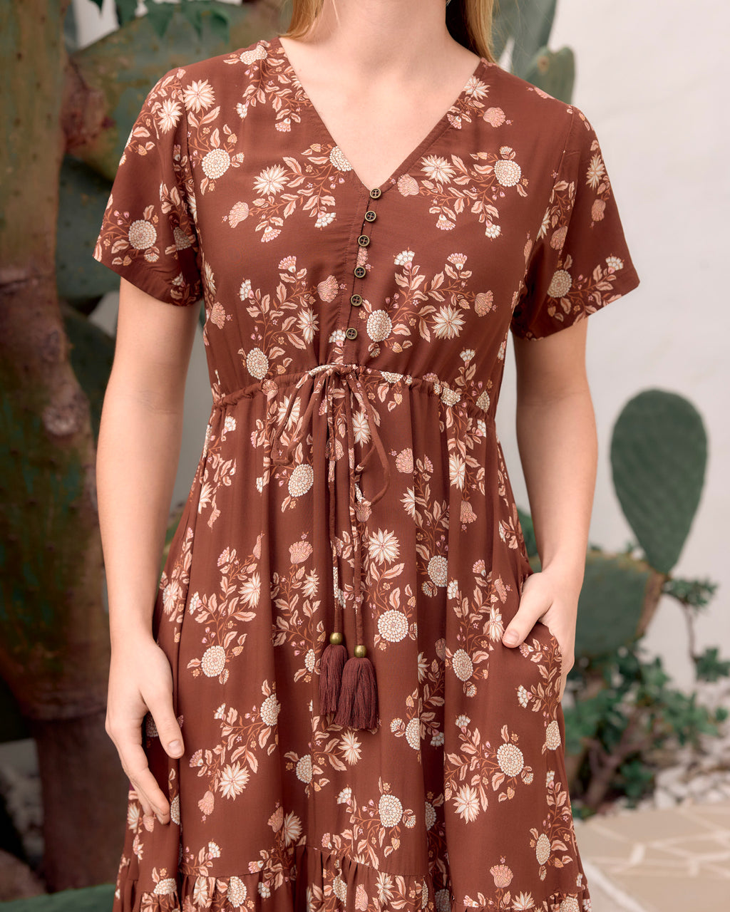 A woman wearing a brown short-sleeve V-neck dress with a beige floral print, button-front bodice, and a drawstring waist with tassel detail, photographed in an outdoor setting with plants in the background.